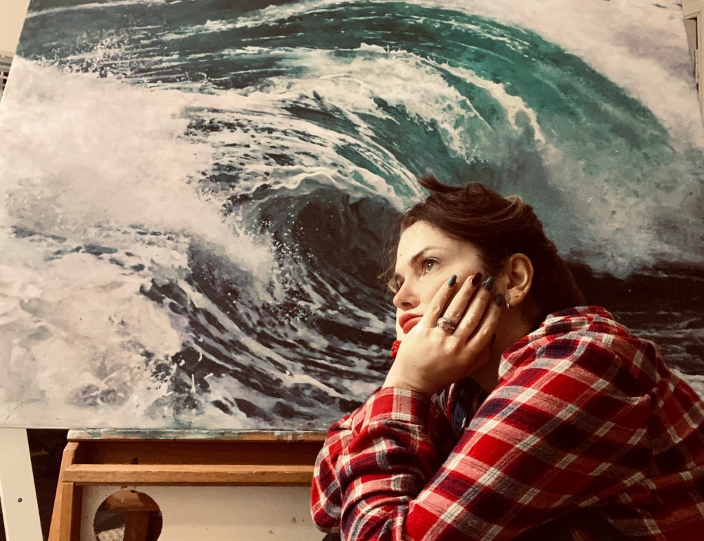 A portrait photo of the artist, a young white woman, sitting pensively in front of a painting of a wave.
