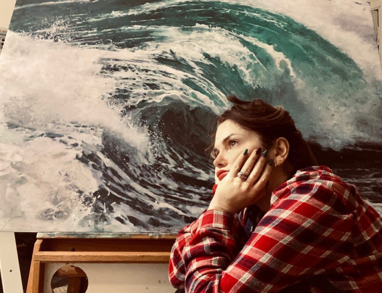 A portrait photo of the artist, a young white woman, sitting pensively in front of a painting of a wave.