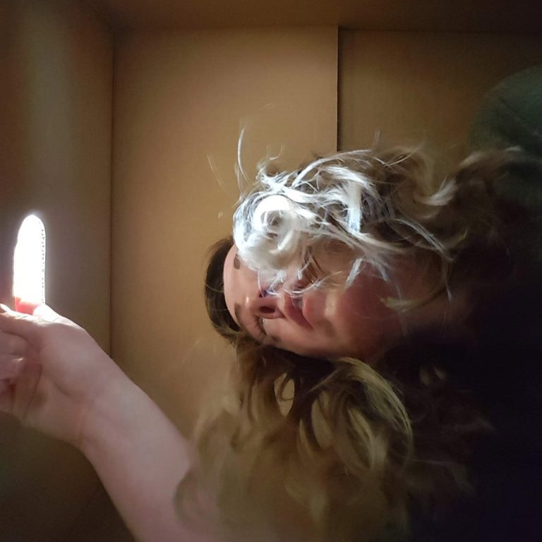 A photo of a young white woman, crouched inside a brown box. The photo is taken from below, and her face is partially obscured by her hair falling over it.