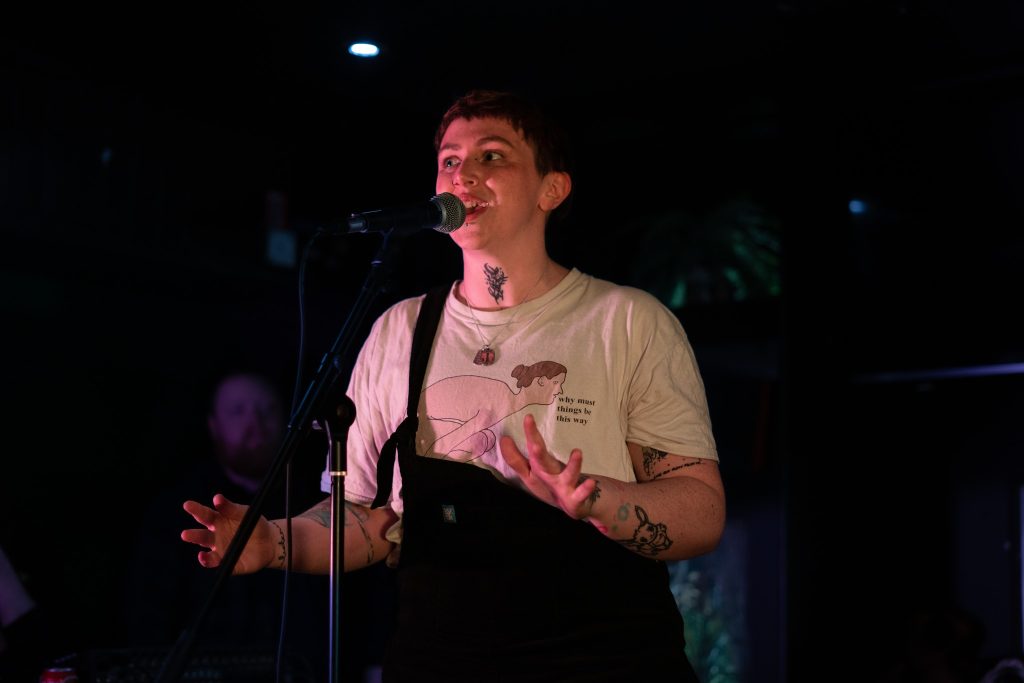 A dramatically-lit photo of Tom, a young white man wearing a white t-shirt, standing in front of a microphone, speaking animatedly.