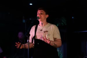 A dramatically-lit photo of Tom, a young white man wearing a white t-shirt, standing in front of a microphone, speaking animatedly.