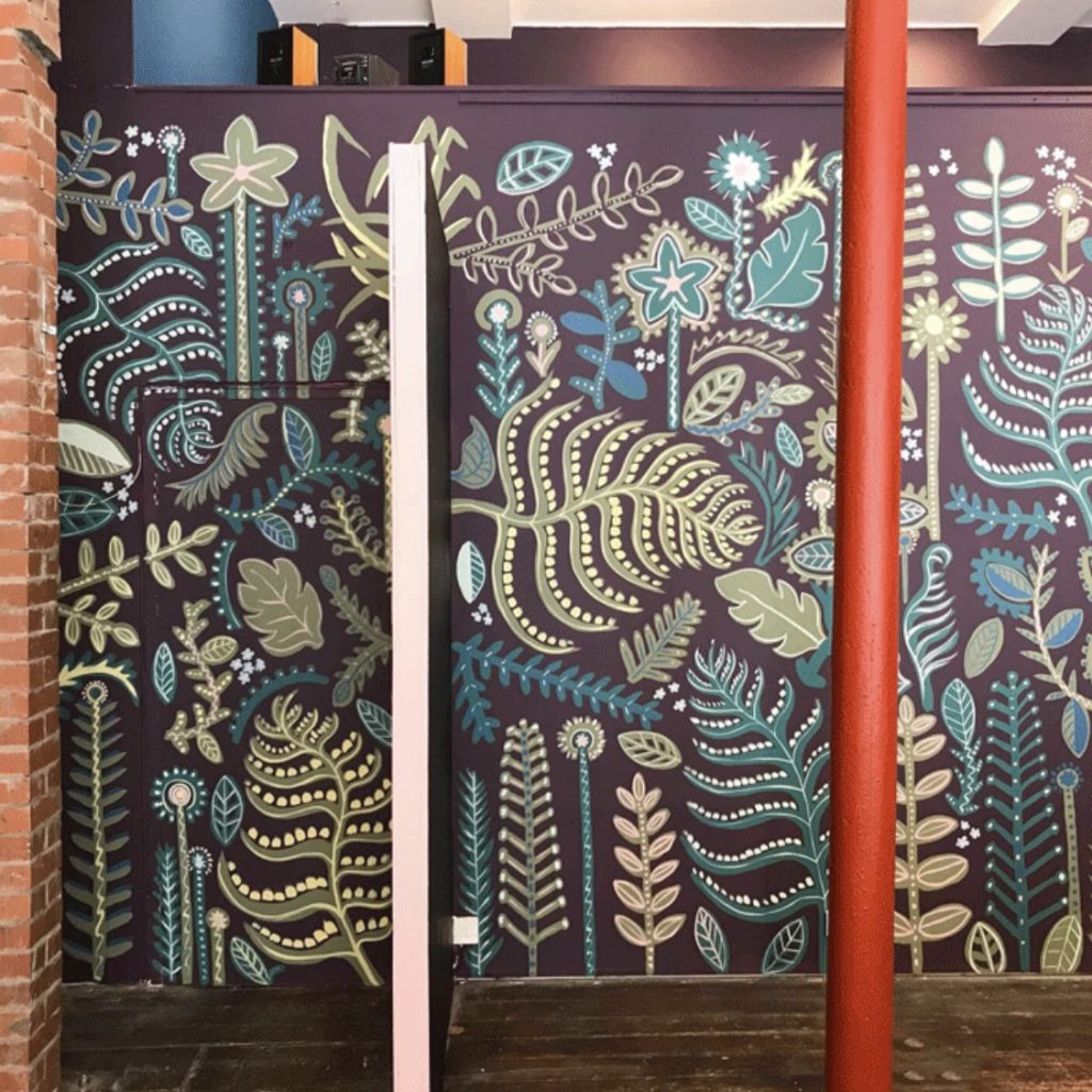 A hand painted wall mural for Refuweegee Charity’s office in Glasgow West End. The large wall is dark purple with lots of leaves and foliage painted over it in greens, blues, pinks and yellows to create a patterned backdrop.
