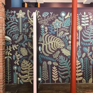 A hand painted wall mural for Refuweegee Charity’s office in Glasgow West End. The large wall is dark purple with lots of leaves and foliage painted over it in greens, blues, pinks and yellows to create a patterned backdrop.