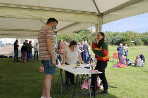 The image shows a brunette female in a colourful jumper talking to a masculine presenting person in a stripy shirt. Between them is a table covered in white paper and art supplies.