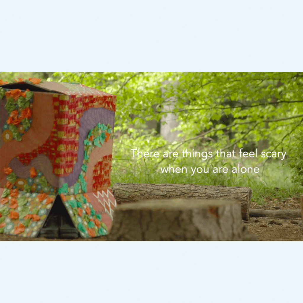 On the left, there is a brightly colored and decorated cardboard box. Black shoes appeared to be coming out underneath the box. On the right, there is the white text 'There are things that feel scary when you are alone'. The image is set in a forest.