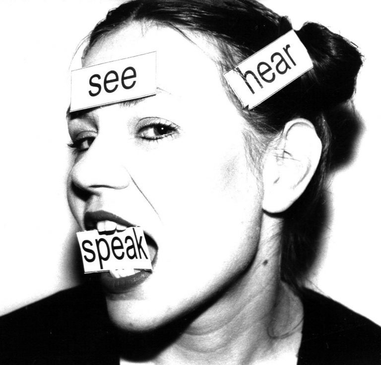 Black & White portrade headshot tilted left with eyes looking into camera with the printed words See, Hear, Speak each physically stuck to forehead, over right ear and held between her teeth in this order.