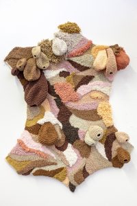 Wall hanging/rug in the shape of a cows hide, made out of different coloured yarns, in skin tones ranging from Dark brown to light cream, warm tan and pinks. Each colour takes a natural shape, resembling different prints from many animals. There are crocheted teats(from cows udders) hanging from the tapestry in the same colours. This piece was made from punch needling techniques, crochet and embroidery.