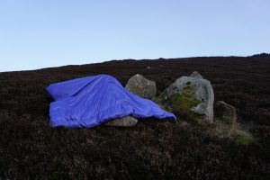 A shiny lilac taffeta (a type of fabric) soft shelled sculpture is draped on top of three mossy small rocks sticking out of a heathery patch on the side of Craig Owl Hill. The soft shelled sculpture is in the shape of a large rectangle, roughly 1.5 meters wide x 3 meters long, the fabric is stuffed and sewn into to make it look like a sleeping bag. The way the soft shelled sculpture lies across the rocks makes it look like an abandoned parachute or sleeping bag. The picture is taken during a Scottish winter sun set and the sky glows a deep purple/blue
