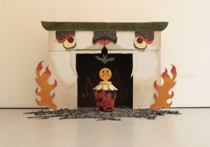 Image is of a uniquely decorated fireplace made out of cardboard, with eyes, a nose, and teeth