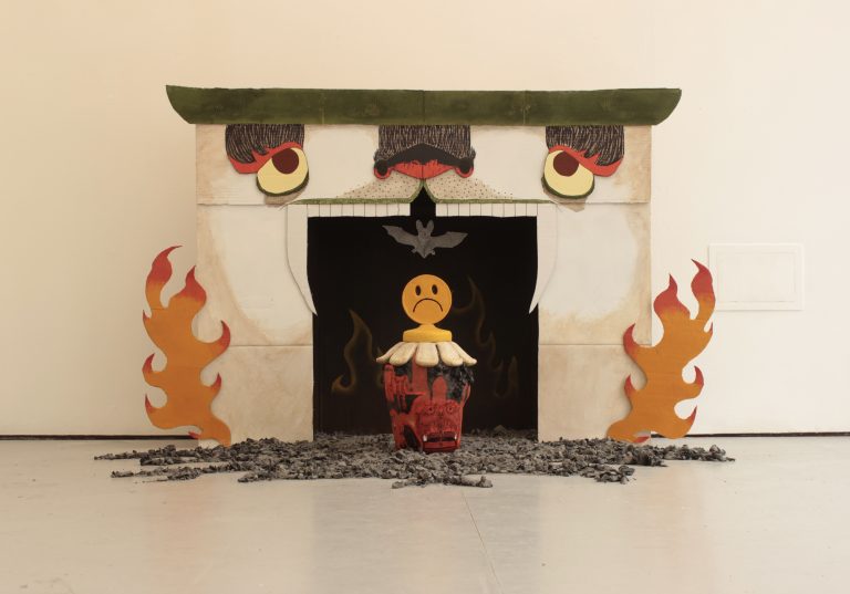 Image is of a uniquely decorated fireplace made out of cardboard, with eyes, a nose, and teeth