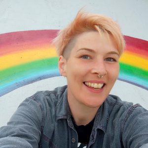 C is a white person with short, peach coloured hair with a shaved undercut. They are shot against a background with a rainbow mural, smiling at the camera.