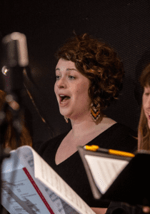 Cathy Phillips Brady, a girl with curly brown hair singing with a folder at BBC Radio Theatre in December 2022