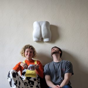 A still from my upcoming short film "Your Ass is Mine."It features two people sitting on the floor against a wall. On the wall is a pair od plaster cast buttocks. The man is looking up at them and the woman is looking out smiling.