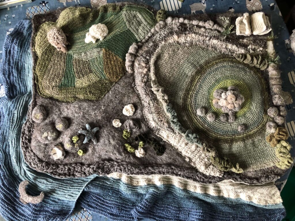 A crocheted landscape with green fields, green and brown circular shapes surrounded by a cream and brown wall. There are star and round shapes in cream, brown and grey. The landscape is surrounded by blue.