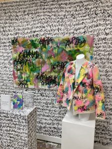 Wall covered in asemic writing, both gray and black. Large layered painting in greens and pinks. Abstract painted, hand sewn jacket on mannequin and three small paintings on top of a plinth also covered in asemic writing.