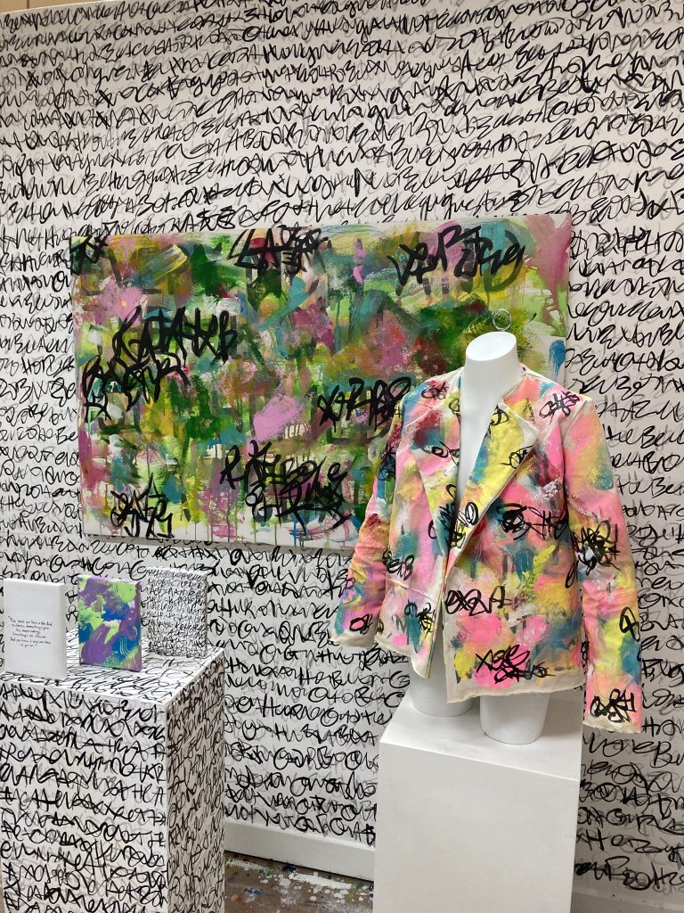 Wall covered in asemic writing, both gray and black. Large layered painting in greens and pinks. Abstract painted, hand sewn jacket on mannequin and three small paintings on top of a plinth also covered in asemic writing.