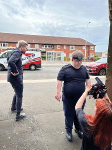 street film shoot with residential building and busy road behind two actors on the pavement while artist crouches holding camera