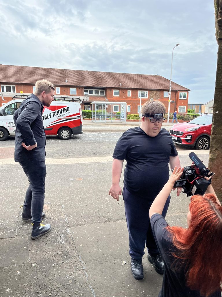 street film shoot with residential building and busy road behind two actors on the pavement while artist crouches holding camera