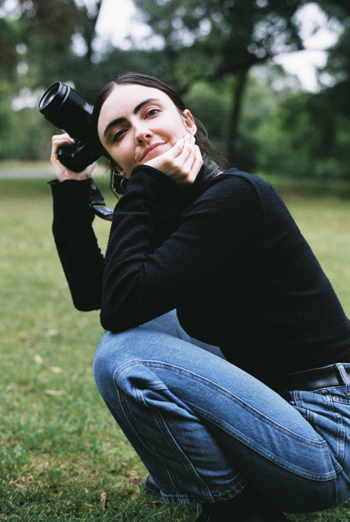 A white woman with dark hair, crouching down and holding a camera.