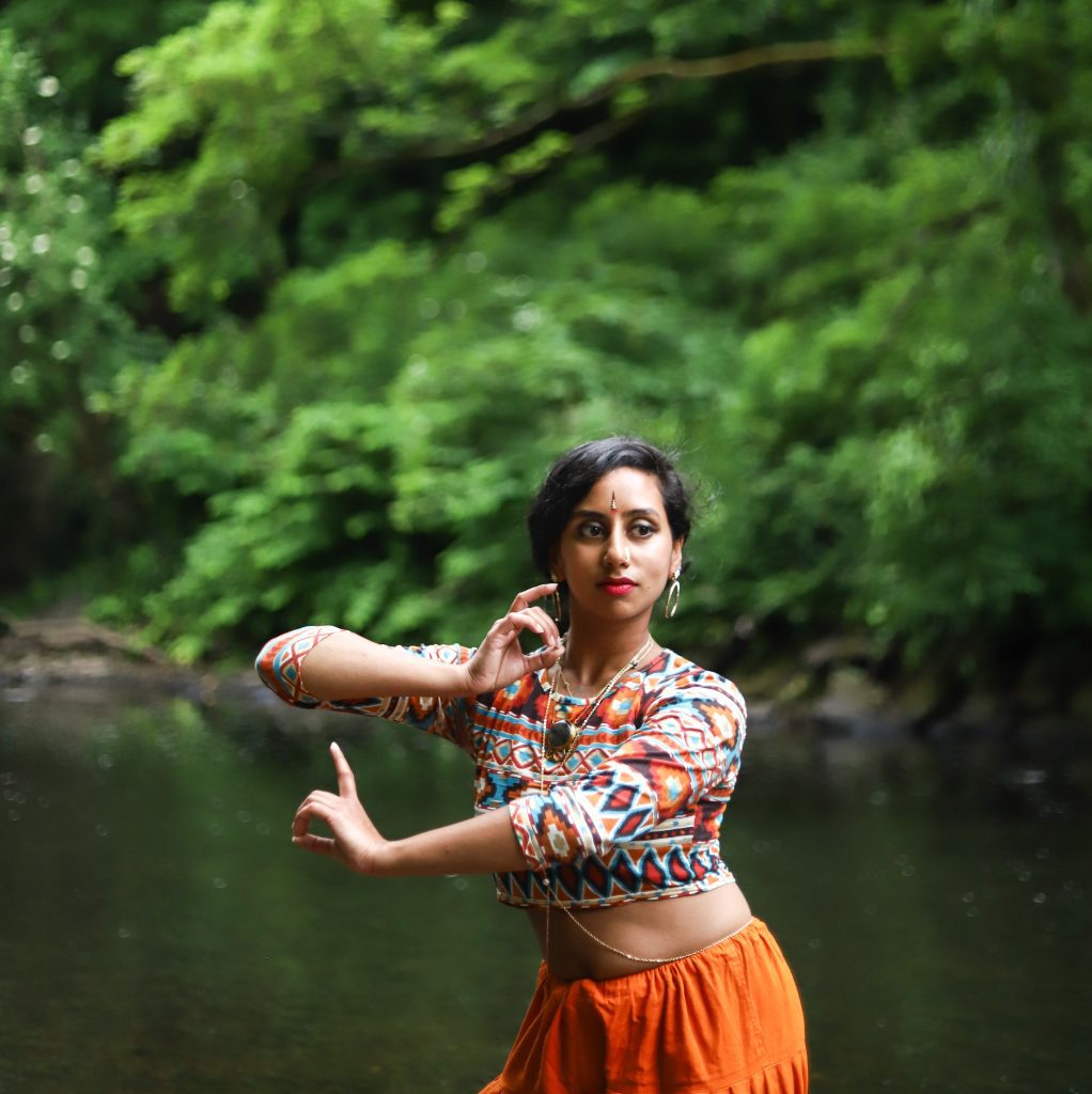 Nikhita stands in nature, before a river, holding a pose commonly seen in Odissi dance