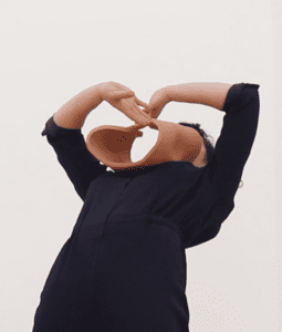 An abstract terracotta object standing on the chest of a dancer in a blue jumpsuit who is back bending whilst reaching her crossed arms towards the object that is resting on her chest