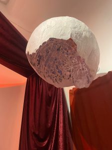 A thin, white, papier-mâché half-dome is suspended from the ceiling. On the inside of the dome there are detailed doodles which light shines through. There are red drapes against white walls in the background.