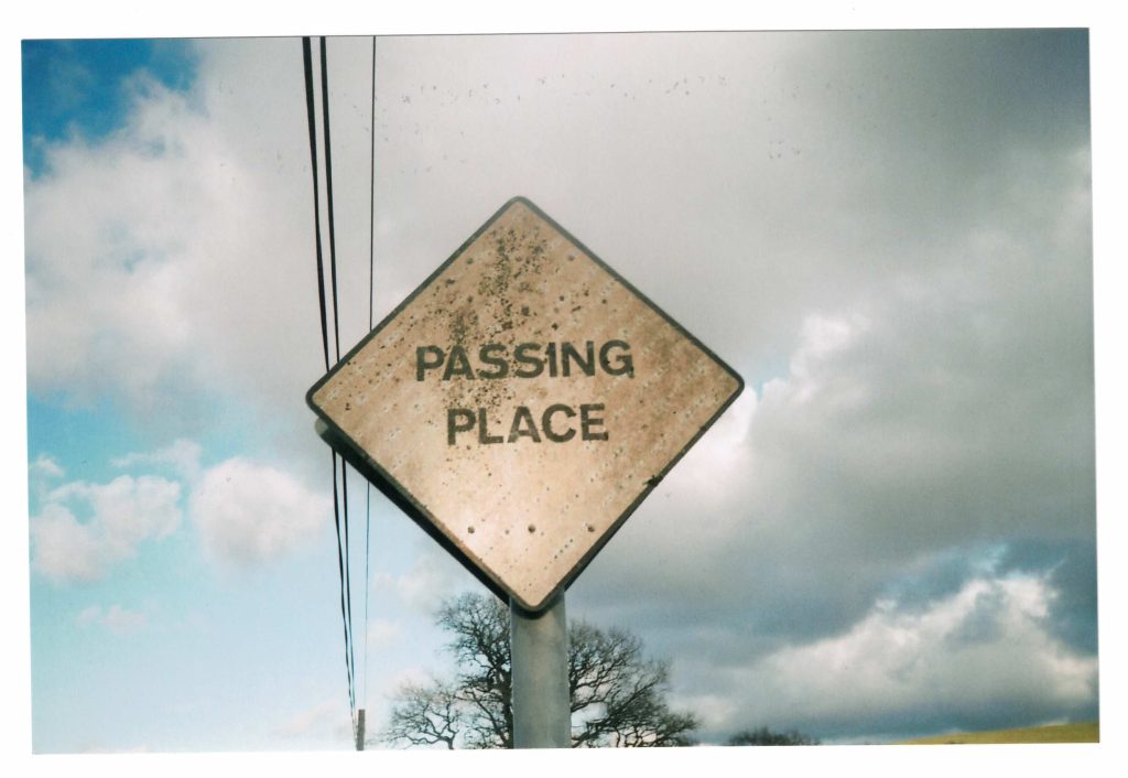 A 4x6 inch printed photograph from 35mm film of a passing place, a diamond-shaped white sign with the words PASSING PLACE. The sky is cloudy and there is a phone line going overhead.