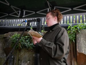 "Eliza stands in the foreground of the image, blond curly hair tied up with a clip. She is wearing a dark green jacket, and a pair of thin silver frames glasses. Eliza is reading from a small publication with a microphone to her mouth. She stands underneath an outside gazebo with a few green wall plants in the background. "