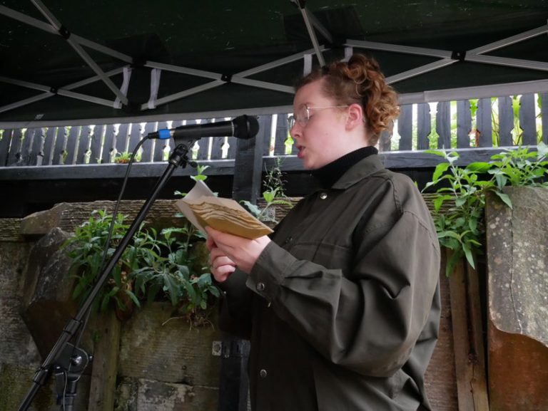 "Eliza stands in the foreground of the image, blond curly hair tied up with a clip. She is wearing a dark green jacket, and a pair of thin silver frames glasses. Eliza is reading from a small publication with a microphone to her mouth. She stands underneath an outside gazebo with a few green wall plants in the background. "