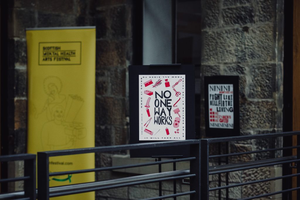 A photo of an A3 risoprinted poster, printed on white paper. In the centre of the poster is a hand written large block text in black which reads ‘No One Way Works’. This quote is continued turning around the outside edges of the page, reading ‘It will take all of us shoving at the thing from all sides to bring it down - Diane di Prima.’ Around the central text is a border of 16 illustrations, which have a thin black outline, and are shaded in three shades of bright red. The illustrations are of: a razer blade; a bunch of Rosemary; a vial (like you would use for Oestrogen); a knotted rope; a pair of scissors; a crowbar; a wheatpaste brush; a balaclava; three round pills with smiley faces on; a spray paint can; a needle; a packet of prescription medications; bolt cutters; a lighter; a dildo; and a brick. The poster is on a poster display board in the CCA, glasgow - the wall, an interior window, and the yellow banner of the Scottish Mental Health Arts Festival are out of focus in the background.