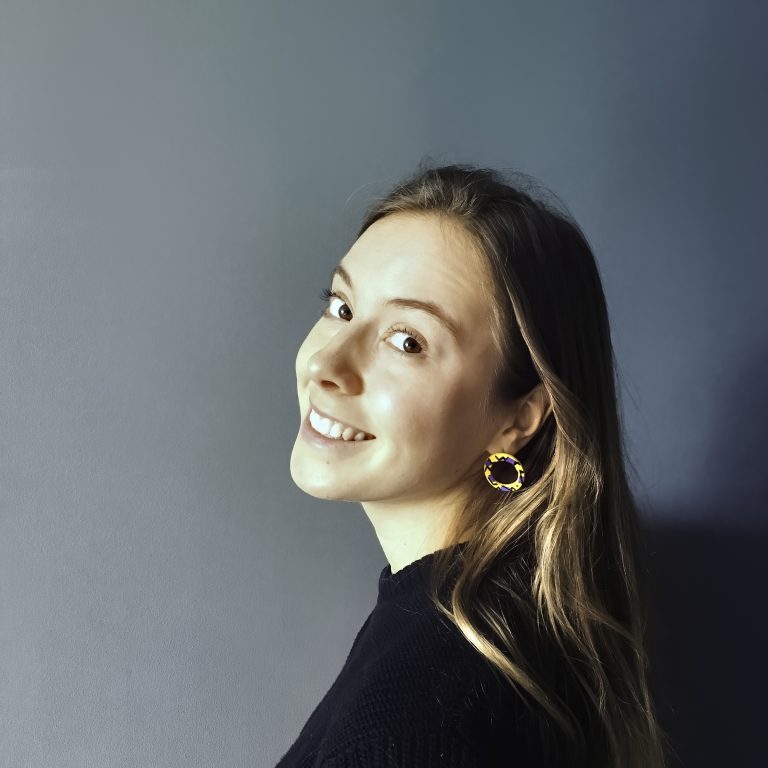 Elspeth Wilson is a white person with long dark blonde hair. They have yellow earrings on and a black jumper and are against a dark grey background. They are smiling and turning to look at the camera.