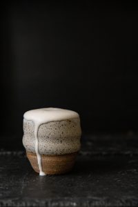 Photo presents white and beige woodfired cup on the black background. The coffee foam is dripping over the rim of the mug all the way down.