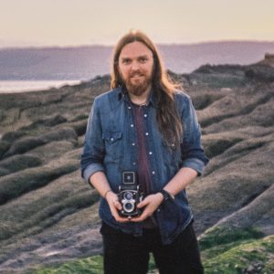 Self-portrait of Alasdair, standing on a beach at sunset holding a vintage TLR camera. Photos shot on 35mm film with Lomo LCA camera.