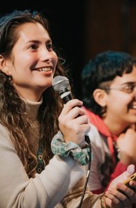 Indigo, a trans feminine fair skin Latina with long curly brown hair wearing a beige turtle neck top holding a mic and smiling. To the right of Indigo, there's a trans masculine person with short hair and glasses wearing a hoodie and smiling.