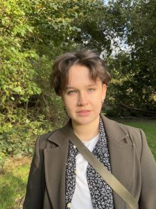 This is a picture of Lyall Tighe smiling at the camera surrounded by trees. They are a white non-binary person with short brown hair and green eyes. They are wearing silver hoop earrings, a long dark green coat, a black floral shirt, a white t-shirt, and a green bag.