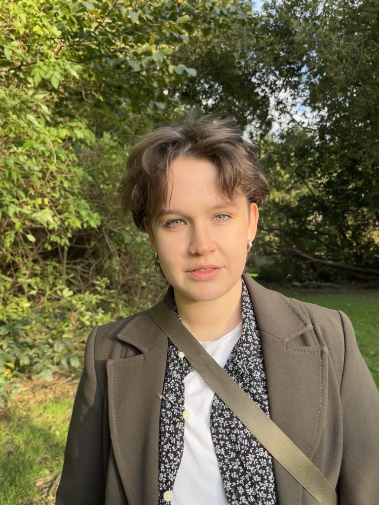This is a picture of Lyall Tighe smiling at the camera surrounded by trees. They are a white non-binary person with short brown hair and green eyes. They are wearing silver hoop earrings, a long dark green coat, a black floral shirt, a white t-shirt, and a green bag.