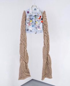 A sleeveless denim jacket covered in badges is hanging from the ceiling. Knitted sleeves have been added to the jacket and are much longer than the jacket itself. The knitting is in brown wool and has random cables designs covering it.