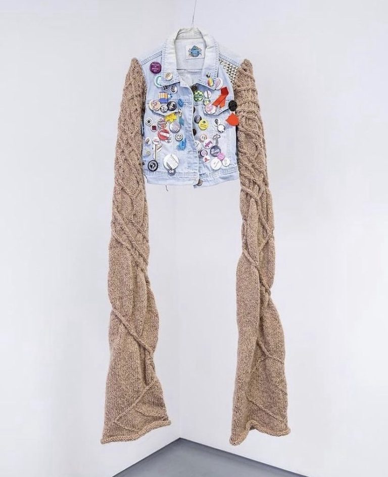 A sleeveless denim jacket covered in badges is hanging from the ceiling. Knitted sleeves have been added to the jacket and are much longer than the jacket itself. The knitting is in brown wool and has random cables designs covering it.