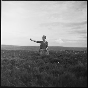 A short-haired person sitting on a chair on a field of grass in the hills. Their right arm is extended and they are looking at their right hand slightly suspiciously.