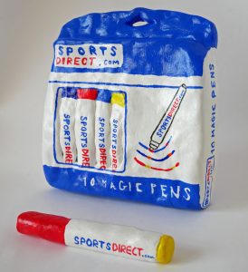 A clay model, decorated in acrylic paint and glazed, of a bag of Sports Direct branded pens with one model lying in front of the bag.