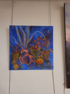 A white wall with a canvas hanging on metal rods suspended from the ceiling The canvas features a painting with a blue background, a pink pig in the background and yellow spider plants.