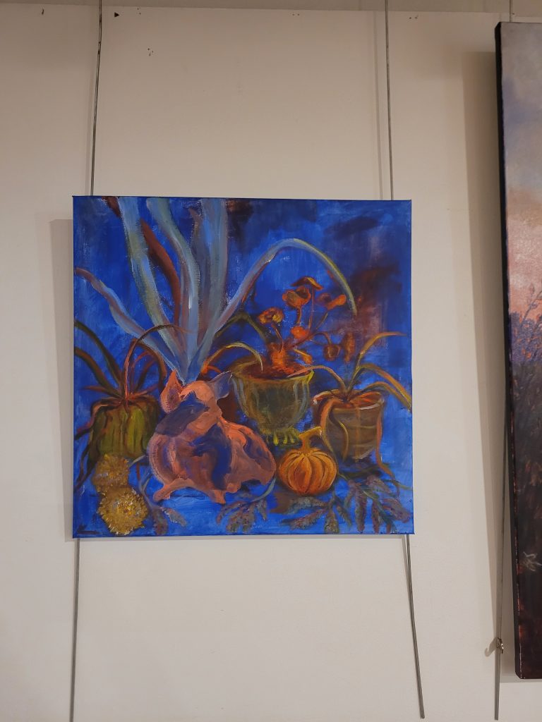 A white wall with a canvas hanging on metal rods suspended from the ceiling The canvas features a painting with a blue background, a pink pig in the background and yellow spider plants.