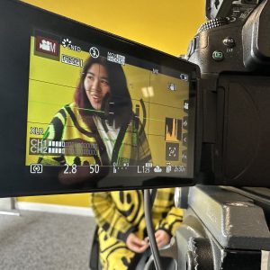 A photo of a video camera display screen with grid lines, pointed at a seated person. The person has shoulder-length purple hair and is looking off to the side. They are wearing a white t-shirt with the text 'Strange', and a yellow cardigan with smiley faces and plaid patterns.