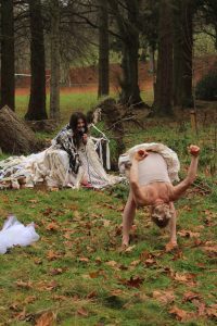 Outside underneath pine trees and upturned roots, one sound artist is in the background wearing a long costume made of hundreds of long strands of white and brown fibres, mimicking the gills and mycelium threads of mushrooms. She is using a microphone and various objects to create sounds which respond to the movements of the Butoh dancer who is bent over, downstage of her, wearing puffy shorts made of the same material.