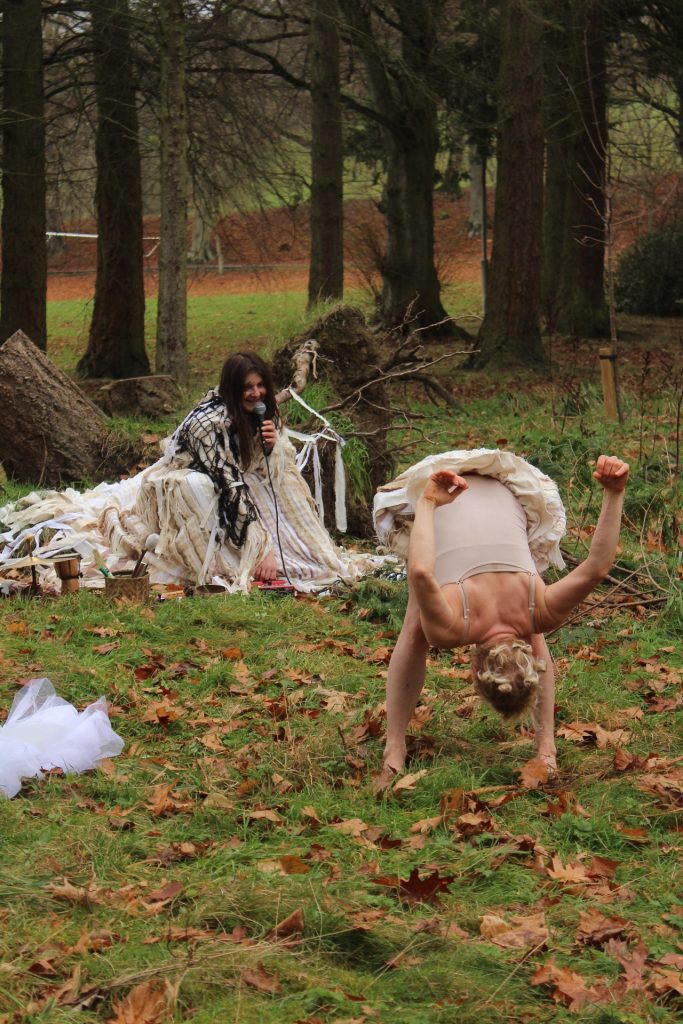 Outside underneath pine trees and upturned roots, one sound artist is in the background wearing a long costume made of hundreds of long strands of white and brown fibres, mimicking the gills and mycelium threads of mushrooms. She is using a microphone and various objects to create sounds which respond to the movements of the Butoh dancer who is bent over, downstage of her, wearing puffy shorts made of the same material.