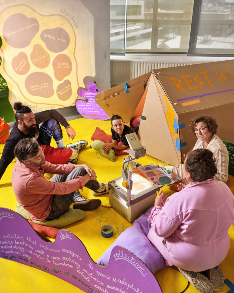Photo of a group of people sitting on the floor in a room full of bright floor mats, and cardboard structures they've built.