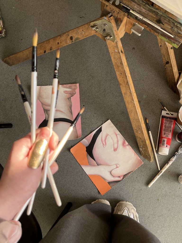 A photo taken from the artist's POV, looking down at paintings on the floor, through paintbrushes held in the left hand. There is an easel peeking into shot. The paintings are of an anonymous woman's body, pulling at non-existent bits of fat.