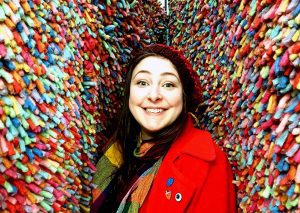 Image shows Lauren Herd smiling and about to laugh, in a red coat and colourful scarf and hat standing against a rainbow rug-like textile background .