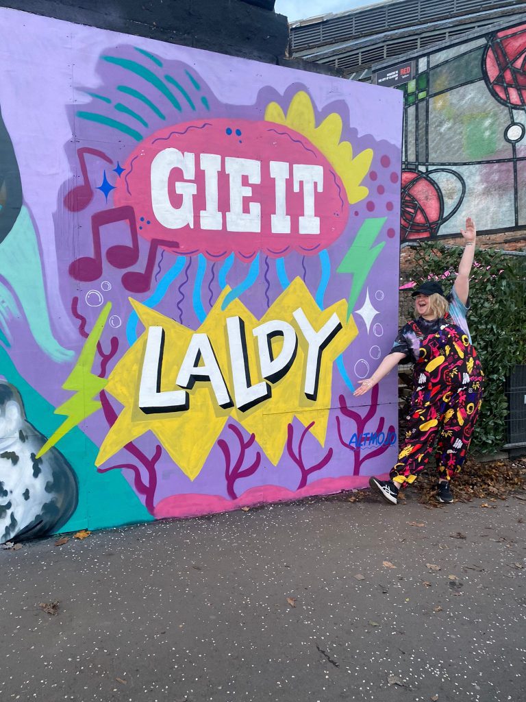 Amy posing infront of a wall mural that says "Gie It Laldy"