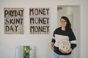 Amanda Lynch standing in the far right of the image with dark shoulder-length hair, glasses, black sweatshirt with white band. Behind figure and to the left are art works, Money money money mixed media work, pay day skint day mixed media art work. Mirror propped on wall bottom left, with cement cast of feet at the bottom